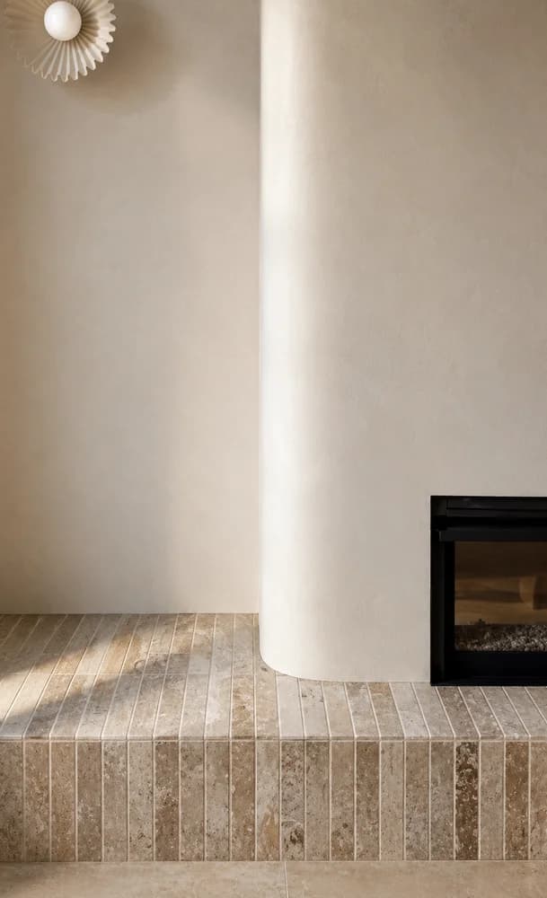 Rounded pale rendered column beside a recessed fireplace with a travertine mosaic tile hearth and a spherical plaster ceiling sconce