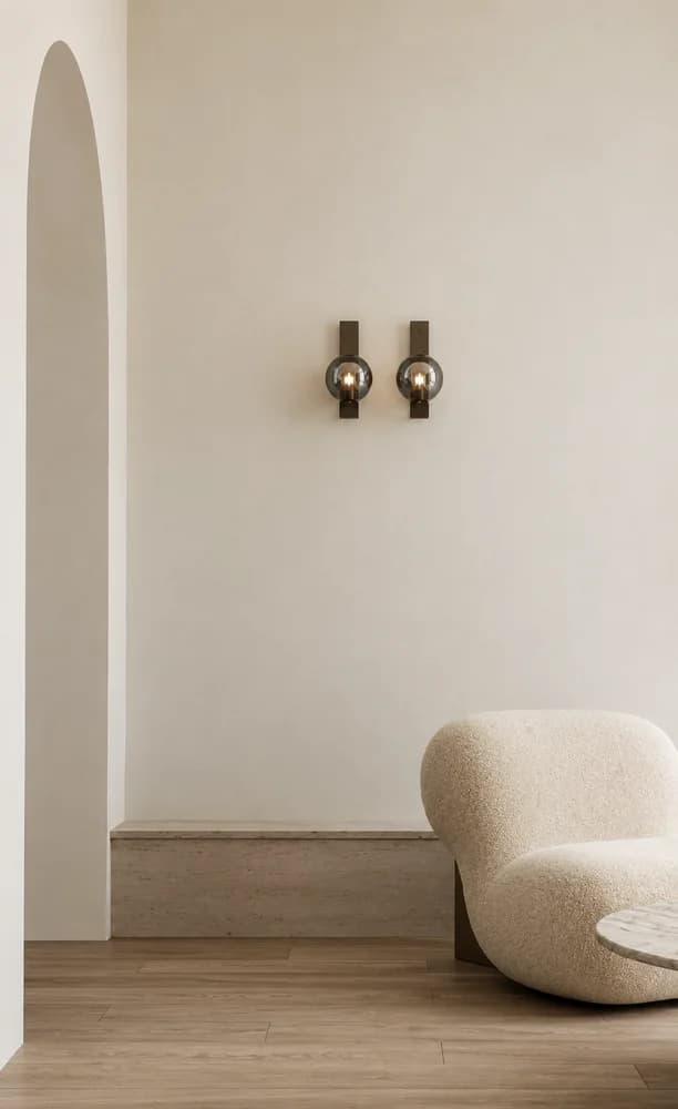 Pale rendered archway opening to a sitting room with warm oak flooring, a bouclé lounge chair and paired smoked glass bronze wall sconces