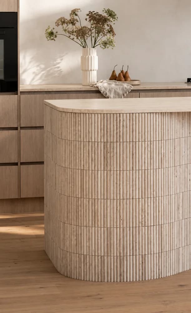 Curved fluted travertine kitchen island with a honed travertine benchtop and warm oak cabinetry behind