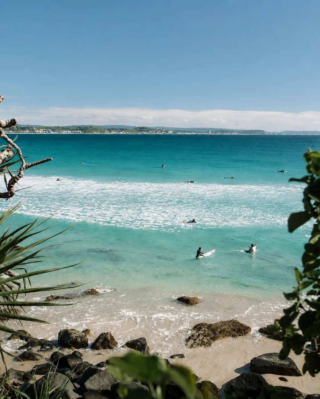 View through coastal foliage to a Byron Bay beach, with surfers sitting on their boards in clear turquoise water and the headland beyond